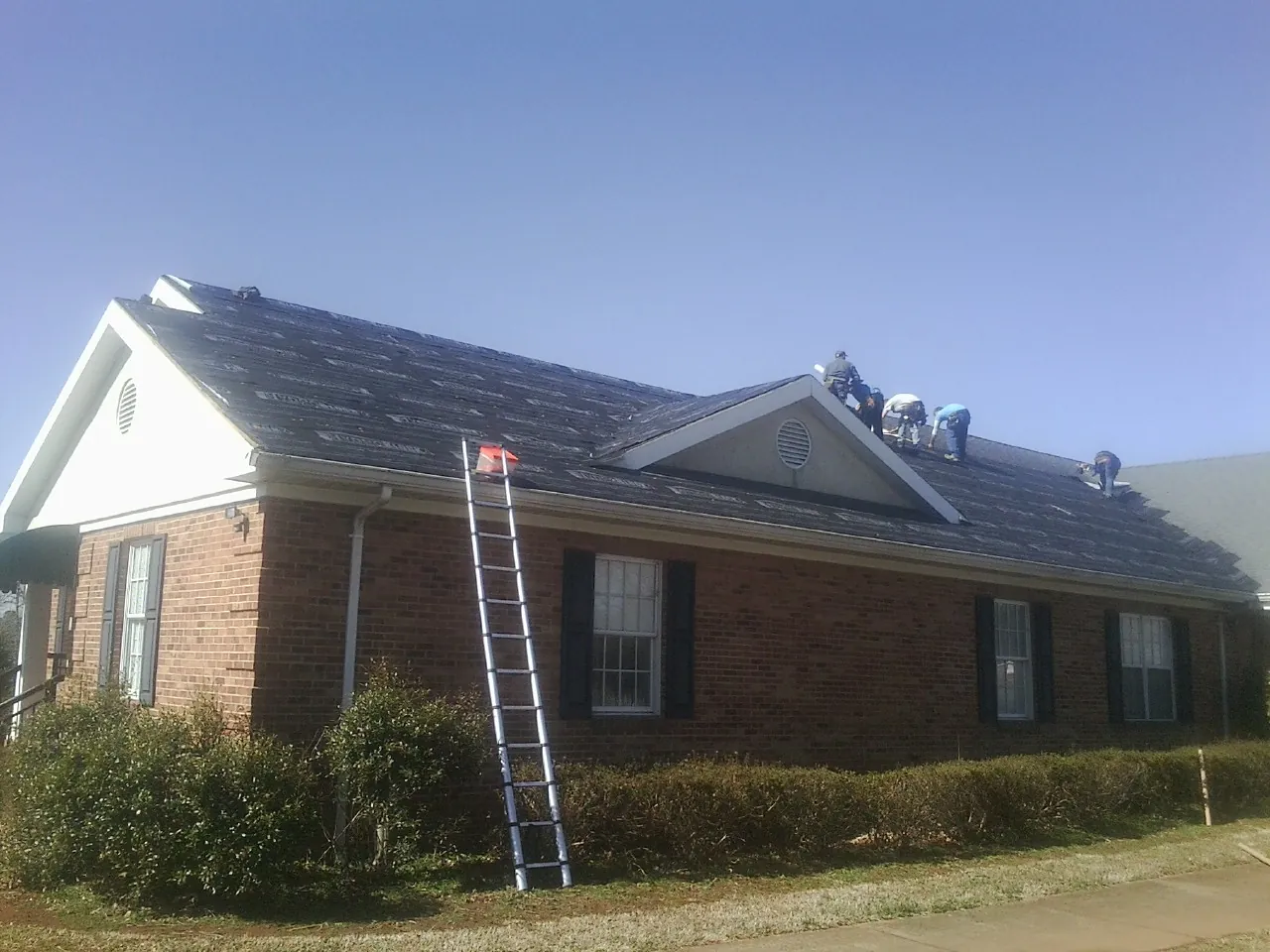 Crew tearing shingles off a brick ranch roof with a ladder set — Emergency Roof Repair work by our crew in Dilliner
