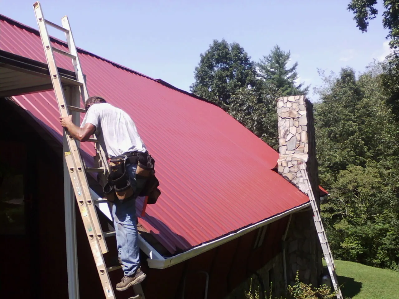 Roofer on a ladder beside a red metal roof and stone chimney — Soffit & Fascia Repair by Roofing Brothers in Dilliner